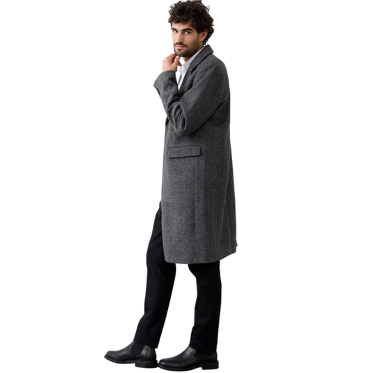 A man wearing a long, gray overcoat with black pants and black leather boots, standing against a white background.