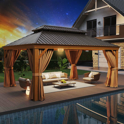 A cedar wood gazebo with a dual-tiered roof and curtains, featuring a sofa set underneath, is placed by a swimming pool near a house at twilight.