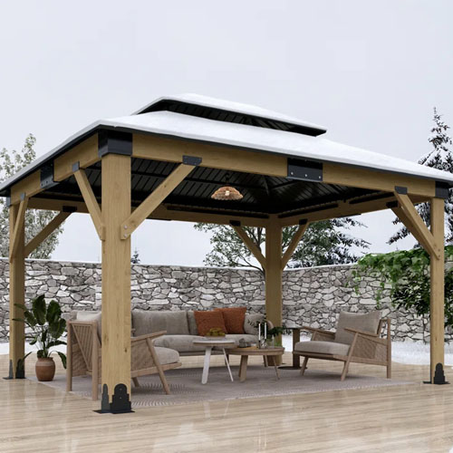 A wooden gazebo with a peaked roof, featuring outdoor furniture including a sofa, an armchair, and a coffee table. The setup is on a wooden deck surrounded by a stone wall and trees.