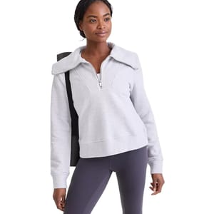 A woman wearing a light gray half-zip sweatshirt with a wide collar and dark leggings carries a yoga mat over her shoulder.