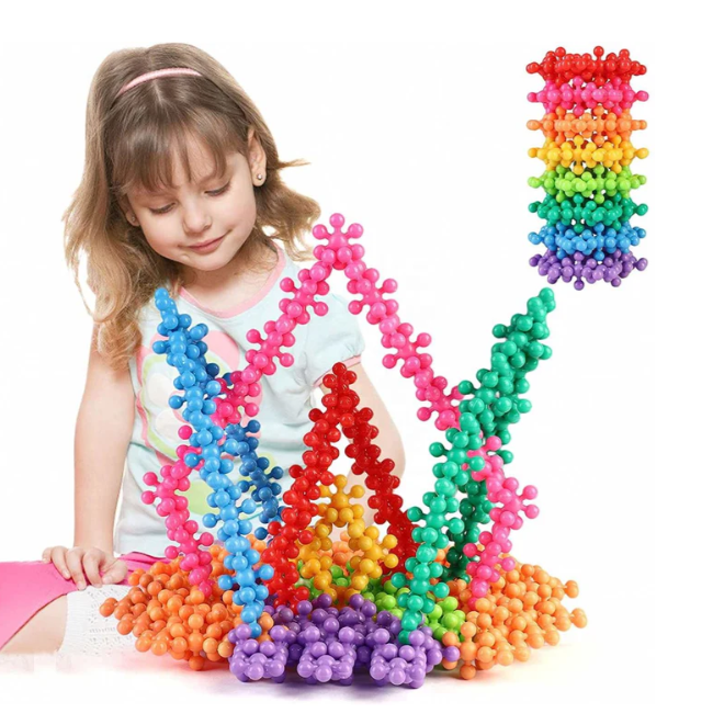A young girl is playing with colorful interlocking plastic building pieces, forming a large Crown and tower-like structure. The building pieces are in various colors, featuring a ball-and-socket design for creative play.