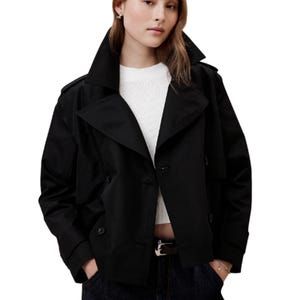 A woman wears a black double-breasted short coat with wide lapels and a cropped design, over a white sweater. The coat has a structured fit with cuffed sleeves and pairs with dark jeans.