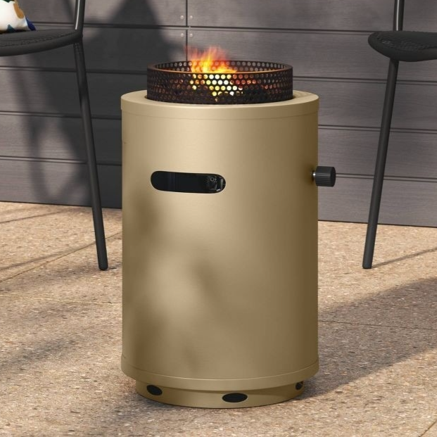 A beige cylindrical outdoor fire pit with a perforated metal top is surrounded by black metal chairs, one with a colorful floral cushion, set against a backdrop of lush green plants.