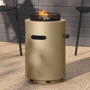 A beige cylindrical outdoor fire pit with a perforated metal top is surrounded by black metal chairs, one with a colorful floral cushion, set against a backdrop of lush green plants.