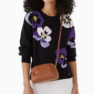 A woman wears a black sweater with large purple flower prints and carries a small tan shoulder bag.