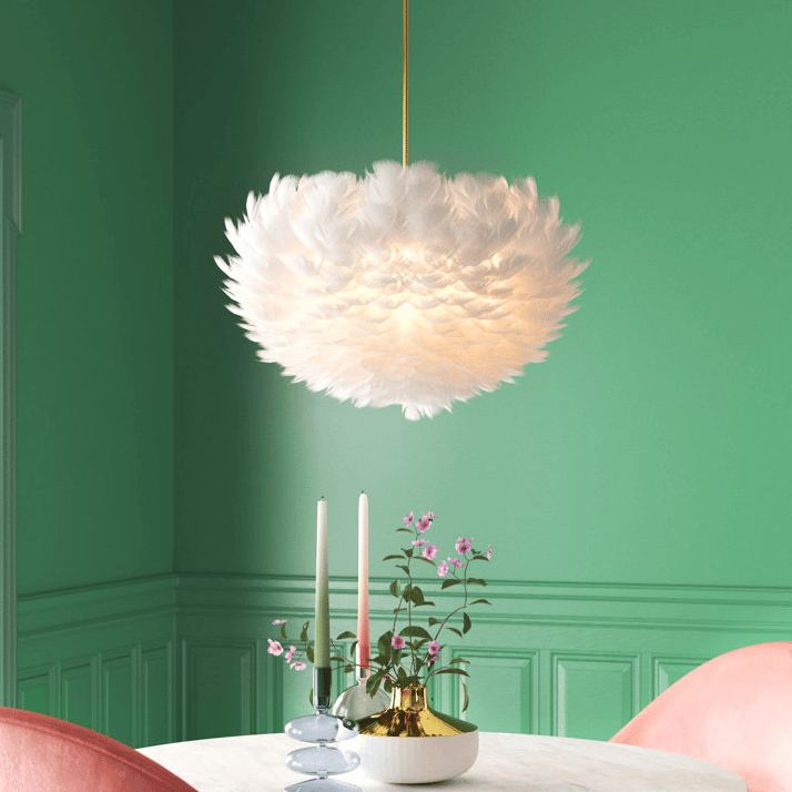 A white feather pendant light hangs above a table with two candles and a gold vase with pink flowers.