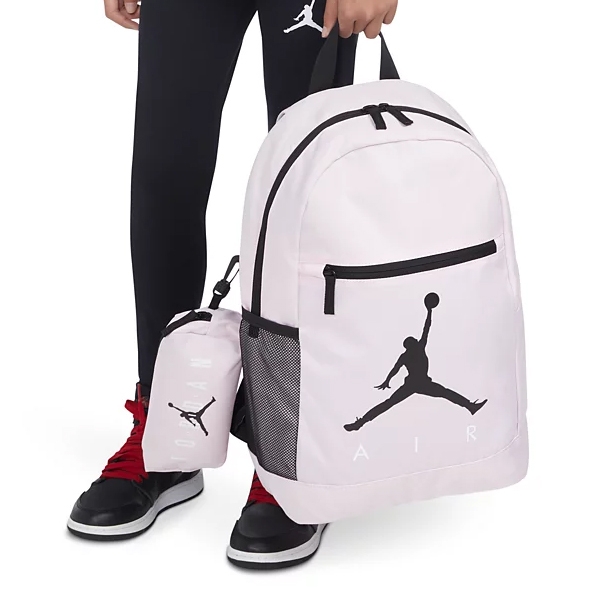 Pink backpack and lunchbox with a black Jumpman logo, held by a person wearing black leggings and red and black sneakers.