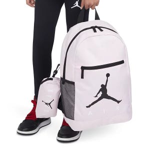 Pink backpack and lunchbox with a black Jumpman logo, held by a person wearing black leggings and red and black sneakers.