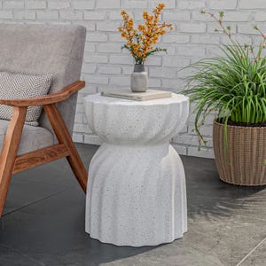 A white, speckled concrete side table with a unique, curvy hourglass shape. It is placed outdoors beside a wooden chair, with a plant and a vase with flowers on top.