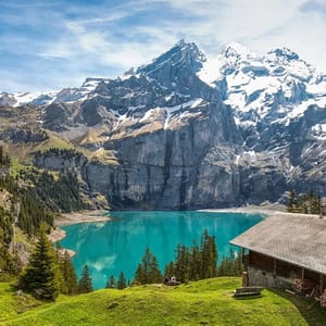 A tranquil alpine scene features a turquoise lake surrounded by snow-capped mountains and lush greenery, with a wooden cabin nearby.