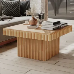 A square wooden coffee table with a fluted design is in a living room setting. It holds stacked books, a small plant in a vase, and a French press.