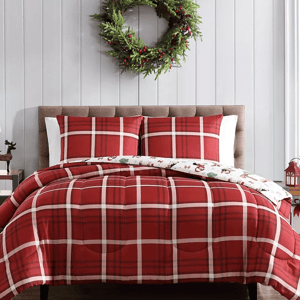 A holiday-themed comforter set featuring a red plaid pattern with reversible dog and festive icon prints, styled on a cushioned headboard bed with matching pillow shams, alongside a decorative wreath above.