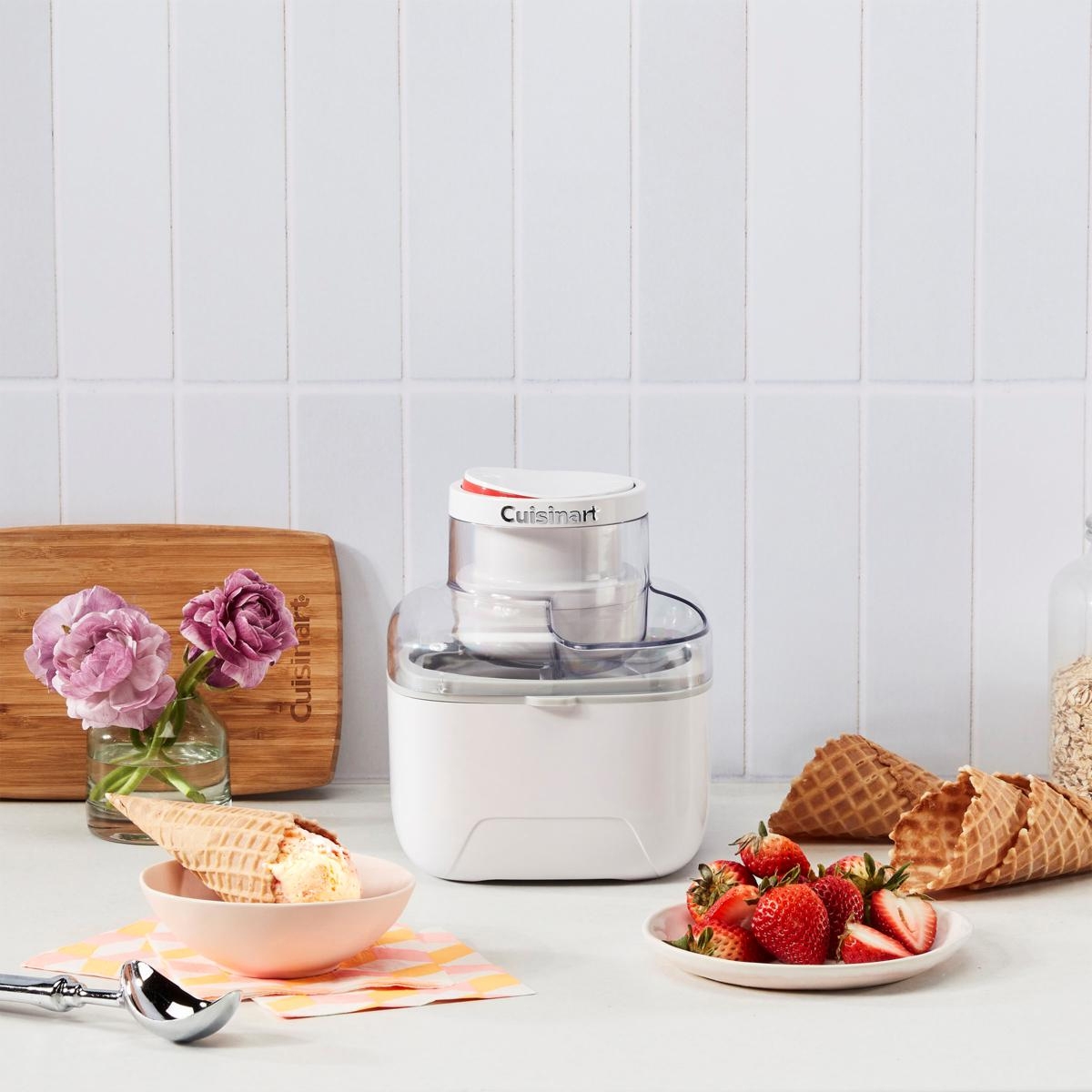 A Cuisinart ice cream maker is displayed next to a bowl of ice cream, waffle cones, and strawberries on a kitchen counter.