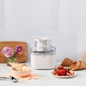 A Cuisinart ice cream maker is displayed next to a bowl of ice cream, waffle cones, and strawberries on a kitchen counter.