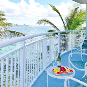 A balcony with ocean views featuring a small table holding a bottle of wine, two glasses, and a platter of fruit, surrounded by palm trees and lounge chairs.