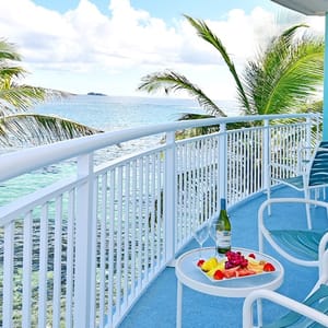 A balcony with ocean views featuring a small table holding a bottle of wine, two glasses, and a platter of fruit, surrounded by palm trees and lounge chairs.