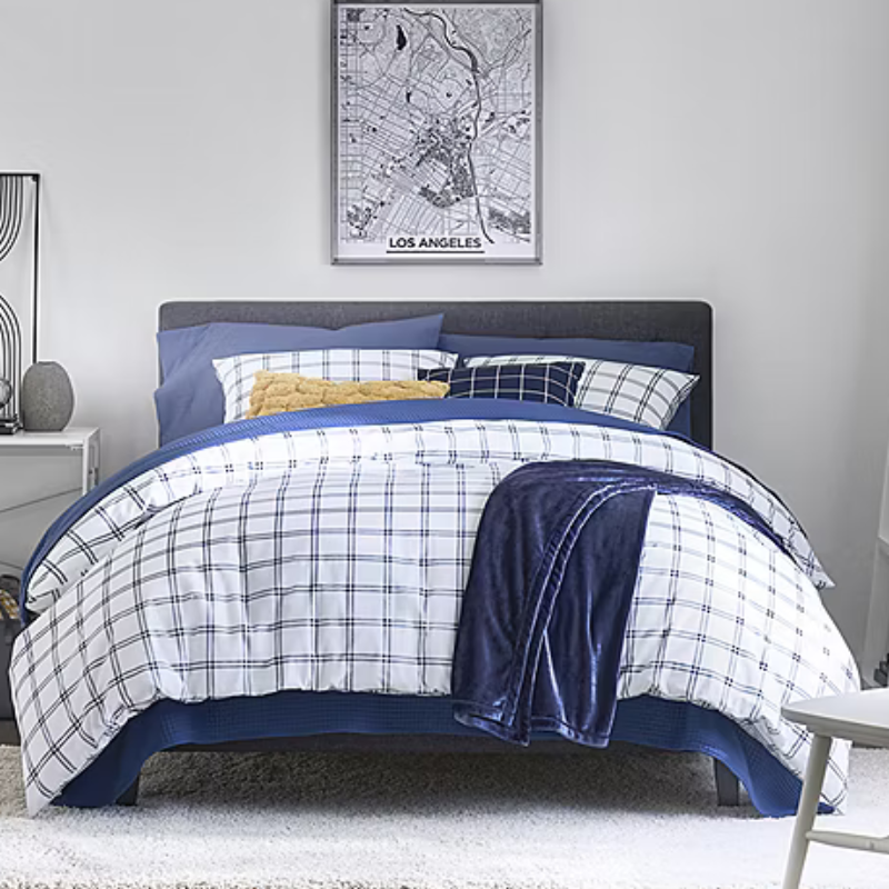 A neatly made bed features a checkered blue-and-white comforter set with multiple pillows, including a yellow accent. A dark blue blanket is draped over the side, with a framed Los Angeles city map on the wall above.