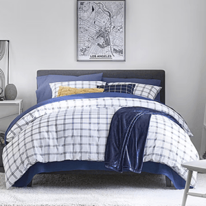 A neatly made bed features a checkered blue-and-white comforter set with multiple pillows, including a yellow accent. A dark blue blanket is draped over the side, with a framed Los Angeles city map on the wall above.