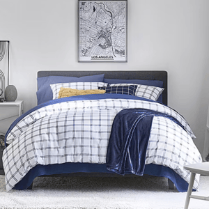A neatly made bed features a checkered blue-and-white comforter set with multiple pillows, including a yellow accent. A dark blue blanket is draped over the side, with a framed Los Angeles city map on the wall above.