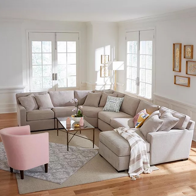 L-shaped sectional sofa in neutral color, pink armchair, two-tiered glass coffee table, decorative throw and cushions, on a beige and gray area rug.