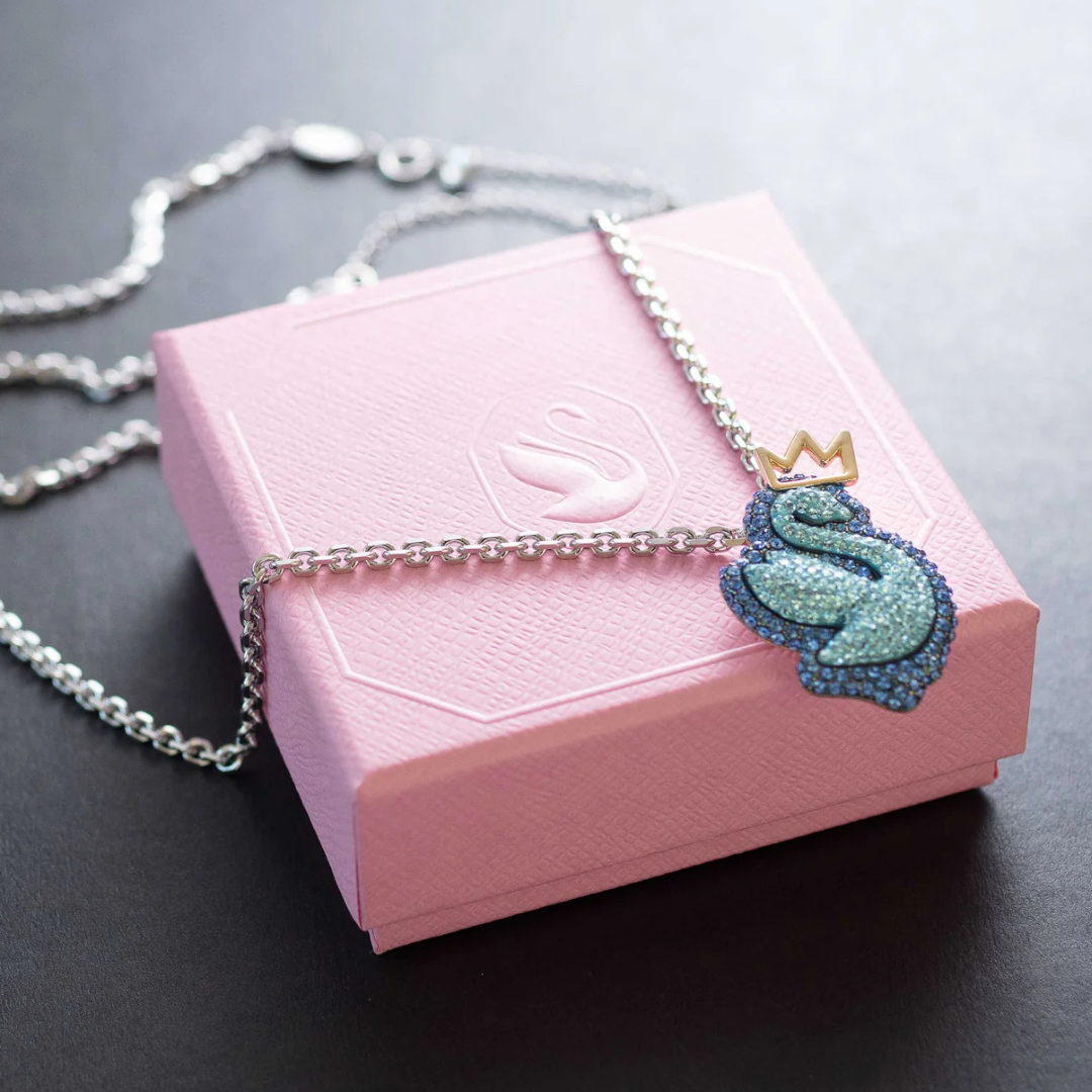 A silver chain necklace with a sparkling swan pendant and a gold crown rests on a pink textured box embossed with a swan logo.