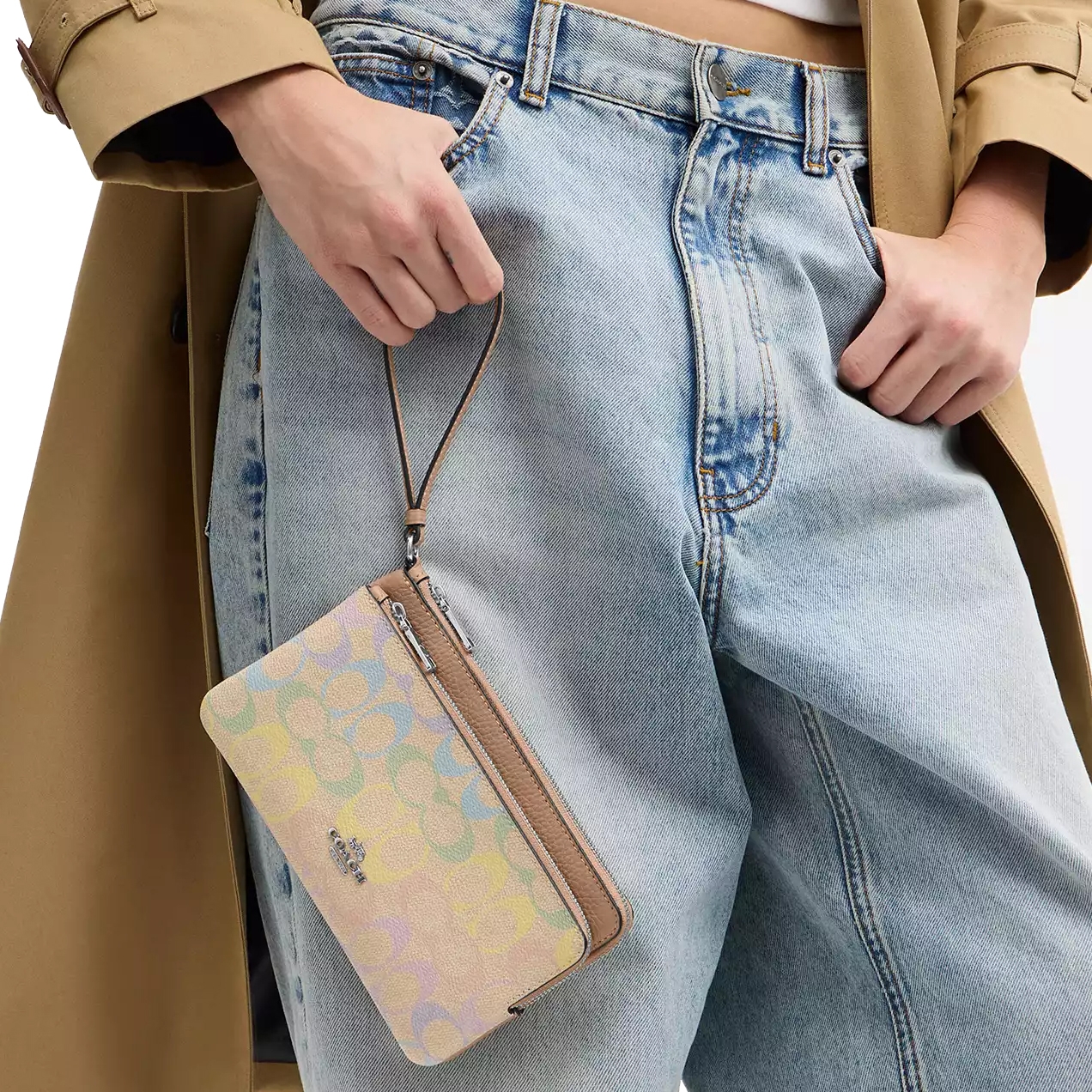 A multicolored wristlet with a subtle pattern is held by a person wearing light blue denim jeans and a beige coat.