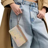 A multicolored wristlet with a subtle pattern is held by a person wearing light blue denim jeans and a beige coat.