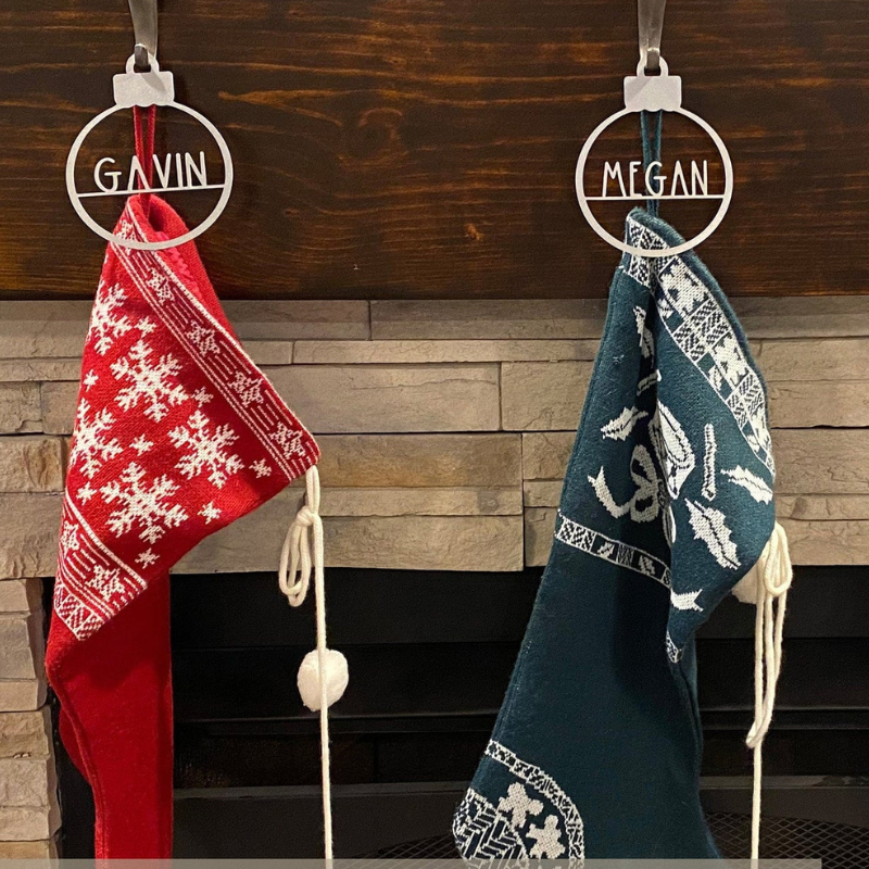 Two Christmas stockings hang from a mantel, one red with white snowflakes labeled \“GAVIN\“ and the other green with festive patterns labeled \“MEGAN\“.