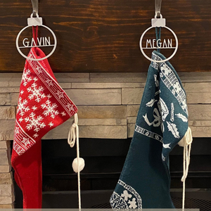 Two Christmas stockings hang from a mantel, one red with white snowflakes labeled \“GAVIN\“ and the other green with festive patterns labeled \“MEGAN\“.