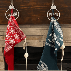 Two Christmas stockings hang from a mantel, one red with white snowflakes labeled \“GAVIN\“ and the other green with festive patterns labeled \“MEGAN\“.