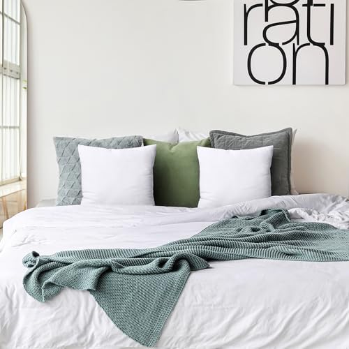 A neatly made bed features white and gray pillows, a central green pillow, and a textured gray blanket. A black and white abstract art piece is mounted on the wall above.