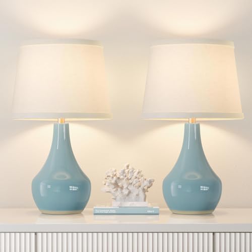 Two blue ceramic teardrop-shaped table lamps with white fabric shades are placed on a white surface, flanking a white decorative coral and a book.