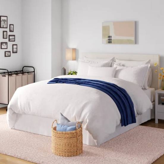 The Wayfair Basics Duvet Set features a simple, elegant design with a white duvet cover complemented by a navy blue throw. It includes matching pillow shams and is placed in a modern bedroom setting with neutral tones and contemporary decor.
