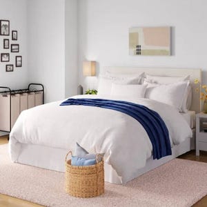 The Wayfair Basics Duvet Set features a simple, elegant design with a white duvet cover complemented by a navy blue throw. It includes matching pillow shams and is placed in a modern bedroom setting with neutral tones and contemporary decor.