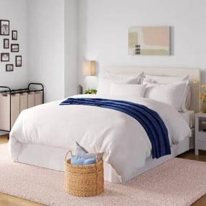 The Wayfair Basics Duvet Set features a simple, elegant design with a white duvet cover complemented by a navy blue throw. It includes matching pillow shams and is placed in a modern bedroom setting with neutral tones and contemporary decor.