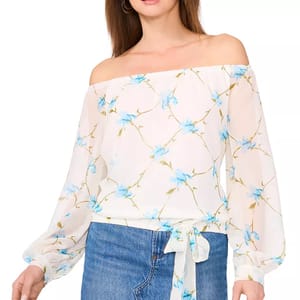 A woman is wearing an off-the-shoulder blouse with long sleeves, featuring a white background and a pattern of blue flowers. She pairs it with blue jeans and hoop earrings.