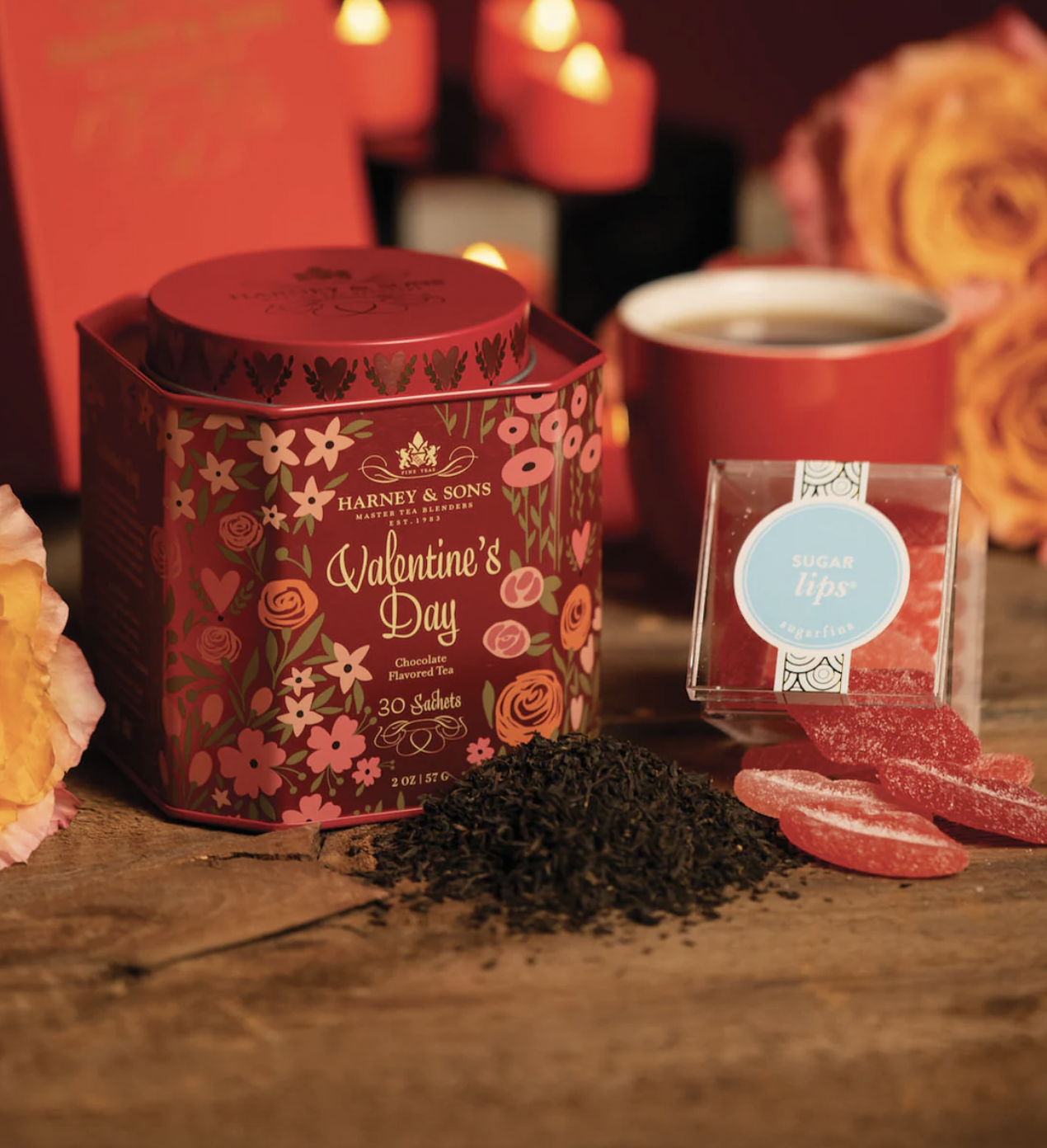 Harney & Sons Valentine's Day Tea Set features a decorative tin of chocolate-flavored tea sachets, displayed alongside a pile of loose tea, a red cup, and Sugarfina \“Sugar Lips\“ gummy candies, all set against a romantic background with candles and roses.