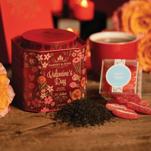 Harney & Sons Valentine's Day Tea Set features a decorative tin of chocolate-flavored tea sachets, displayed alongside a pile of loose tea, a red cup, and Sugarfina \“Sugar Lips\“ gummy candies, all set against a romantic background with candles and roses.