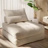 A beige, plush floor chair with a matching pillow is set in a room with a potted plant and a side table.