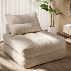A beige, plush floor chair with a matching pillow is set in a room with a potted plant and a side table.