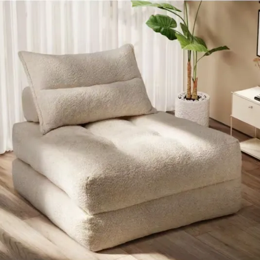 A beige, plush floor chair with a matching pillow is set in a room with a potted plant and a side table.