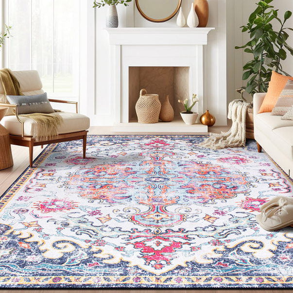 A colorful, machine-washable rug measuring 8' x 10' features an ornate design with blue, pink, and orange patterns, set in a bright and airy living room with a white fireplace, modern furniture, and potted plants.