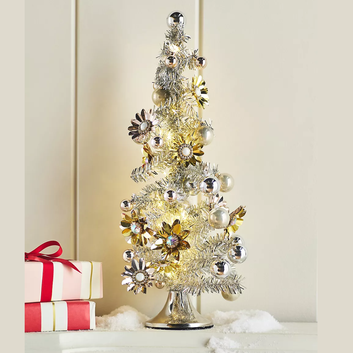 A small silver tabletop Christmas tree decorated with metallic flowers and baubles, illuminated with warm lights, sits on a surface with artificial snow and two wrapped gifts beside it.