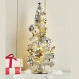 A small silver tabletop Christmas tree decorated with metallic flowers and baubles, illuminated with warm lights, sits on a surface with artificial snow and two wrapped gifts beside it.