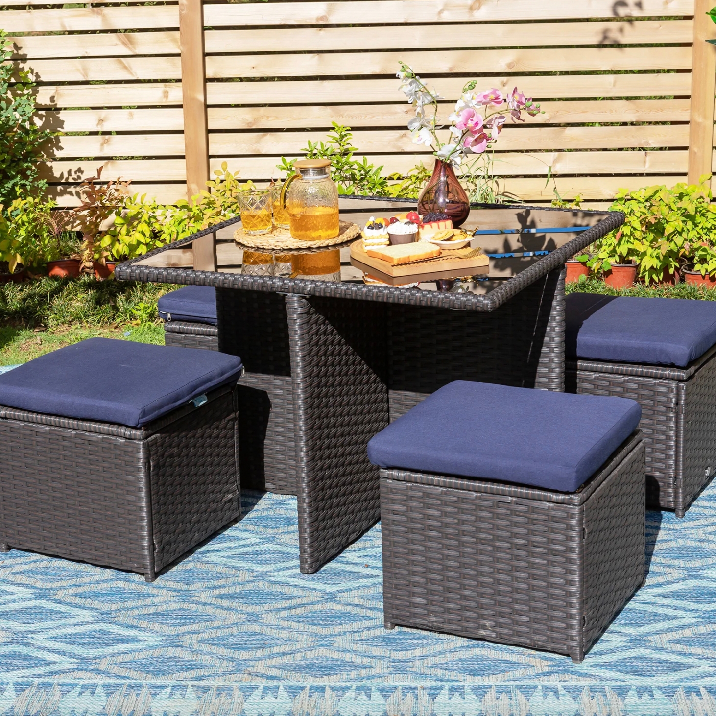 Outdoor patio set with a square glass-top table and four cushioned stools, featuring a woven rattan design. A pitcher of juice, glasses, a tray of snacks, and a flower vase are on the table.
