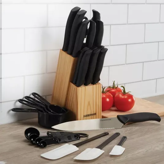 A knife set in a wooden block with black handles, accompanied by measuring cups, spoons, two white spatulas, a peeler, and a cutting board with tomatoes.