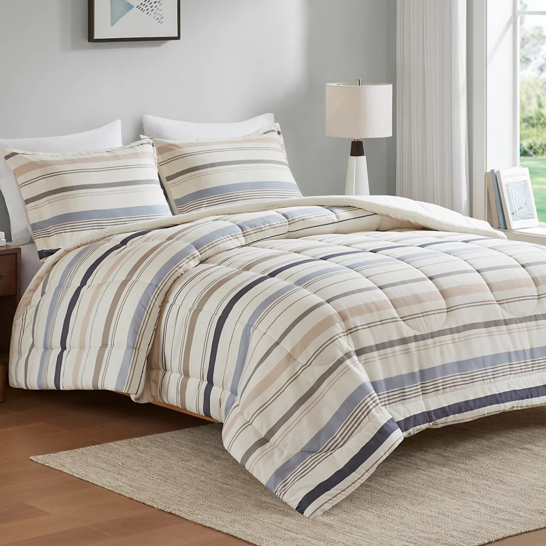 A bed is made with a striped comforter and matching pillow shams in shades of beige, gray, and navy. The room features a nightstand with a lamp and a window with white curtains.