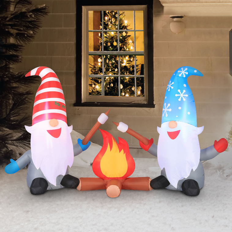 Two inflatable gnomes with tall hats—one red and white striped, the other blue with snowflakes—sit around a faux campfire. Behind them is a window with a decorated Christmas tree.