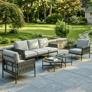 Outdoor patio set with a stone-paved area, including a three-seater sofa, two chairs with rope detailing, and two round coffee tables. Surrounded by a landscaped garden with lush greenery and a stone pathway.