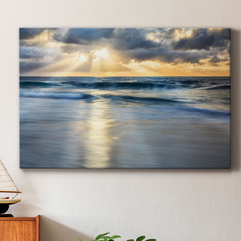 A 36\“ x 24\“ canvas depicts a serene ocean scene with waves gently rolling onto the shore, under a dramatic sky with clouds and beams of sunlight breaking through. A small model sailboat is on a nearby wooden surface, adding a touch of nautical charm.